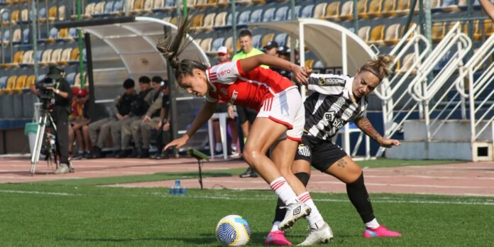 Two female soccer players battle for the ball on the field, one in red and white and the other in black-and-white stripes, with benches and cameras visible in the background.