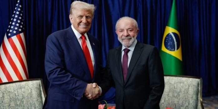 Two men in dark suits shake hands and smile in front of American and Brazilian flags at a formal event.