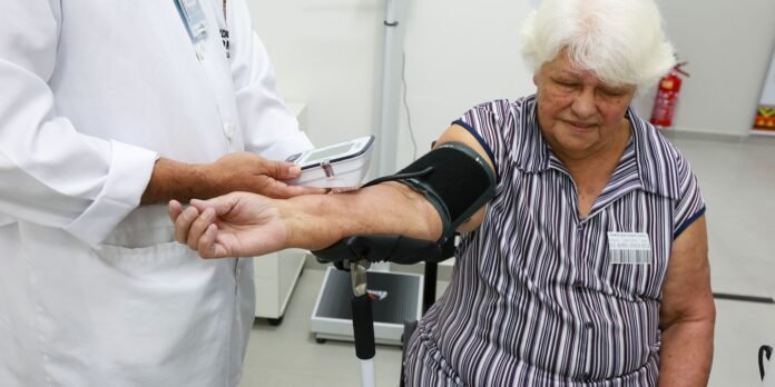 Healthcare worker measures an elderly woman’s blood pressure using a cuff on her upper arm. In the background, a monitor is attached to the cuff.