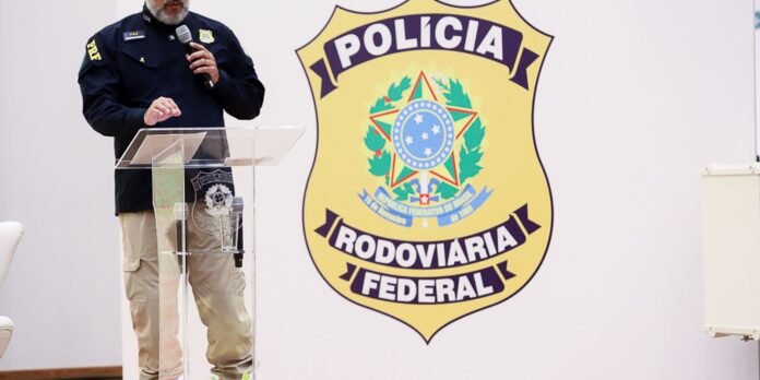 A police officer in a dark uniform speaks at a glass podium, with a large Polícia Rodoviária Federal badge banner behind him.