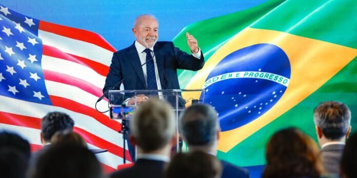 Man in a suit giving a speech at a clear podium with the U.S. and Brazil flags as a backdrop