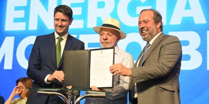 Three men in suits on stage presenting a framed document; one holds it open for display while others smile, with a blue 'ENERGIA' backdrop behind them.