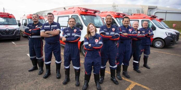 EMS team of nine paramedics poses with arms crossed in front of a row of red-and-white ambulances.