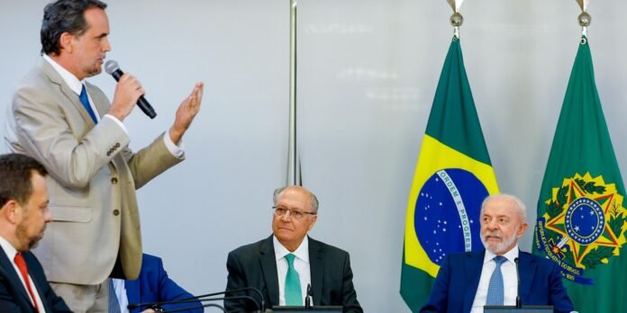 Man in a beige suit speaks into a microphone while gesturing, at a formal meeting with two flags in the background (Brazil and another green flag).