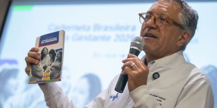 Male speaker in white coat holding a microphone and a booklet titled 'Caderneta Brasileira da Gestante' during a presentation.