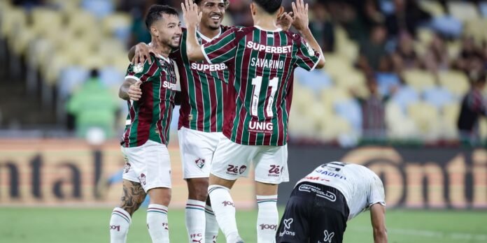 Soccer players in maroon-green striped jerseys celebrate a goal on the field, with arms raised and teammates embracing.