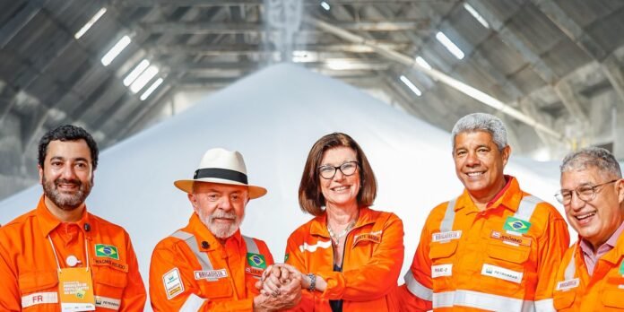 Five professionals in orange safety uniforms stand together in a large industrial space, hands joined in the center and smiling at the camera.