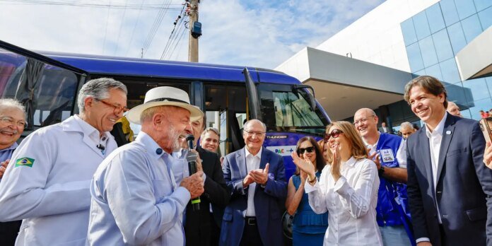 Group of people smiling and clapping around a blue bus during an outdoor event, man with microphone in white hat speaking.