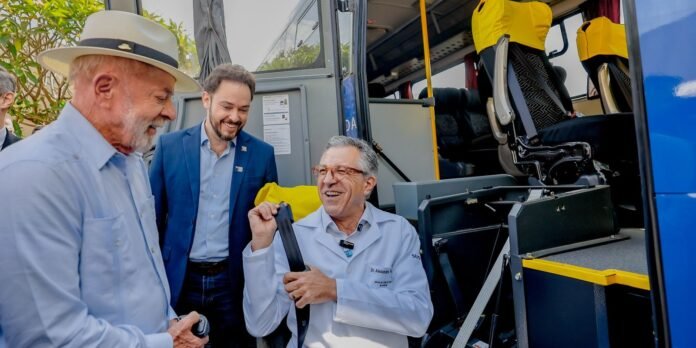 Three men outside a blue bus; a doctor in a white coat shows a seat belt while others smile.