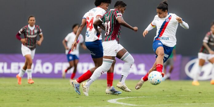 Players in white and striped jerseys chase a soccer ball during a match on a green field.