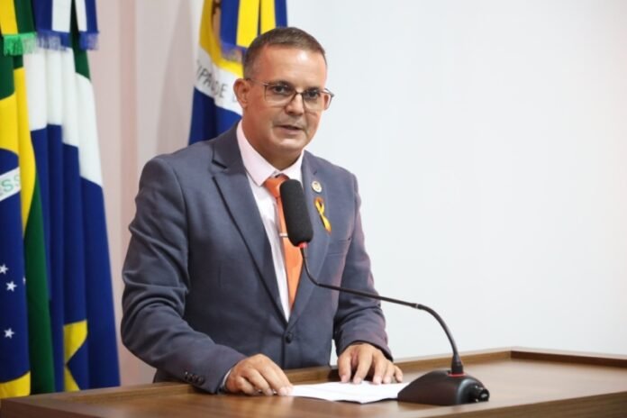 Man in a dark suit at a wooden podium giving a speech, with a yellow ribbon pin and flags in the background.