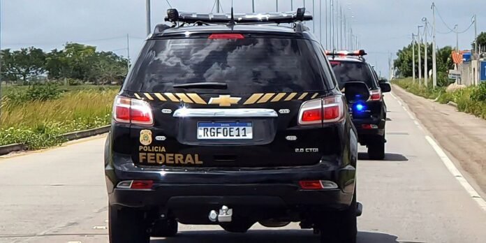 Black Polícia Federal SUV with yellow diagonal stripes, license plate RGFOE11, trailing a police vehicle with blue lights on a rural road.