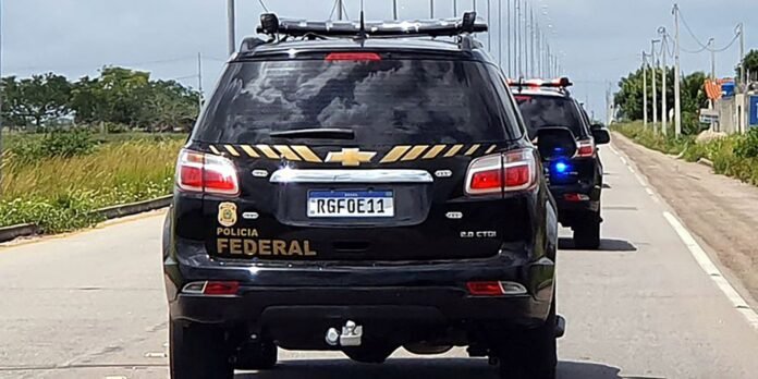 Black Polícia Federal SUV with yellow diagonal stripes and roof rack driving on a rural road, another patrol car behind it likely in pursuit/escort.