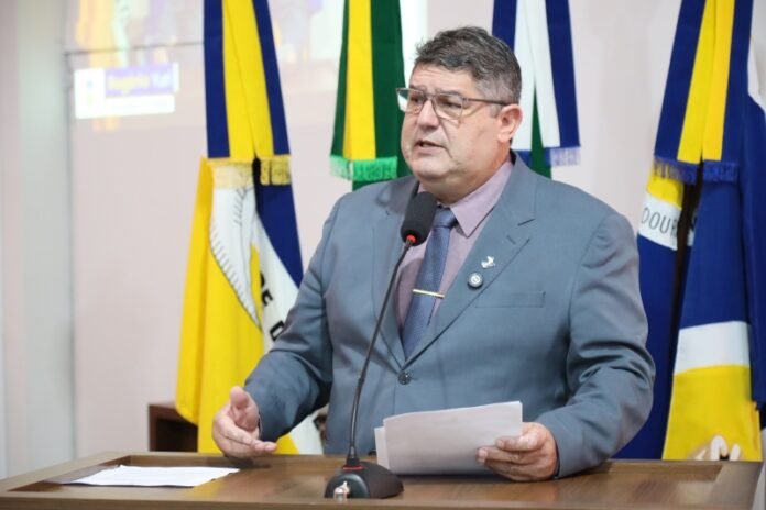 Man in a gray suit speaks at a podium with papers and a microphone, colorful flags behind him.