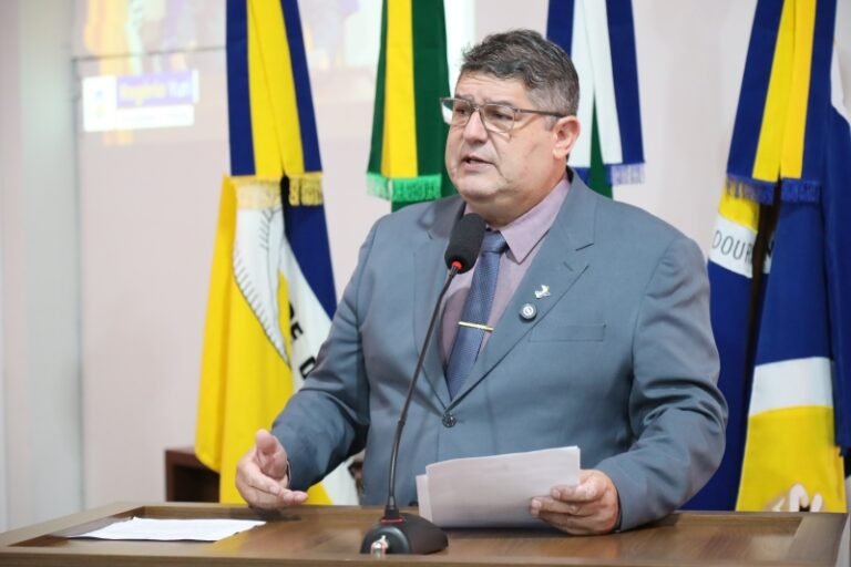 Man in a gray suit speaks at a podium with papers and a microphone, colorful flags behind him.