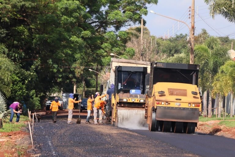 Construction crew in orange shirts paving a street with an asphalt paver and a nearby dump truck.