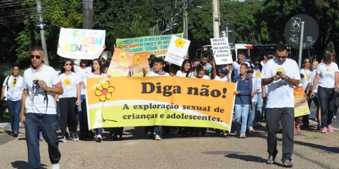 Group of protesters marching on a sunny street, holding a large orange banner that reads “Diga não! A exploração sexual de crianças e adolescentes.” with flower illustration, while others carry signs in Portuguese advocating against abuse of minors.