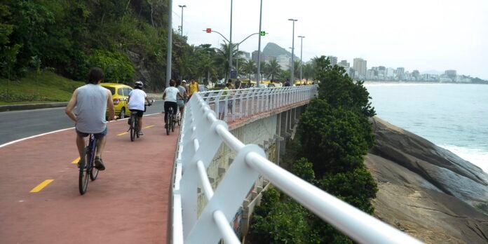 Cyclists ride a red bike path along the coast, beside a white railing, with palm trees, rocky shore, and a distant city skyline.