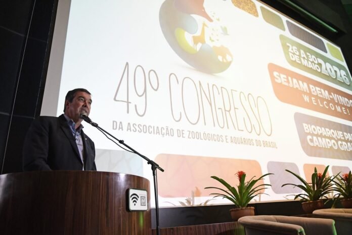 Homem em palanque de madeira fala ao microfone durante congresso, com tela gigante ao fundo exibindo '49º CONGRESSO DA ASSOCIAÇÃO DE ZOOLÓGICOS E AQUÁRIOS DO BRASIL'