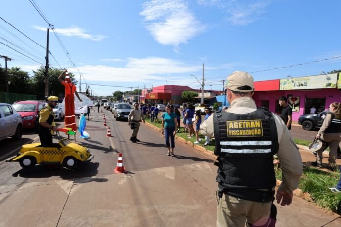 Street parade scene with performers on a yellow taxi-cart and an orange stilt-walker, crowd watching along a lined avenue under a blue sky, cones marking the route, and security personnel overseeing the event.