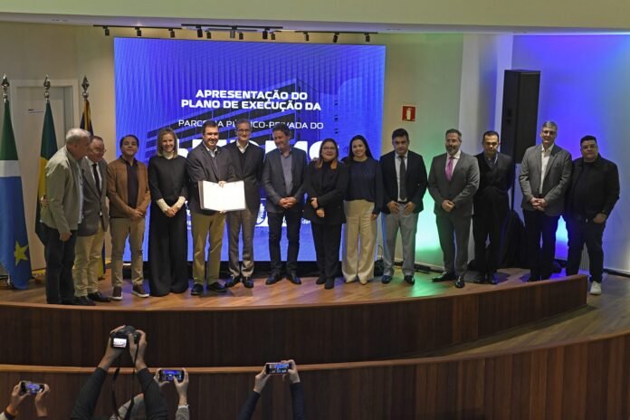 Group of professionals on a stage posing for a photo during a formal ceremony, with a blue screen backdrop behind them and flags on the left.