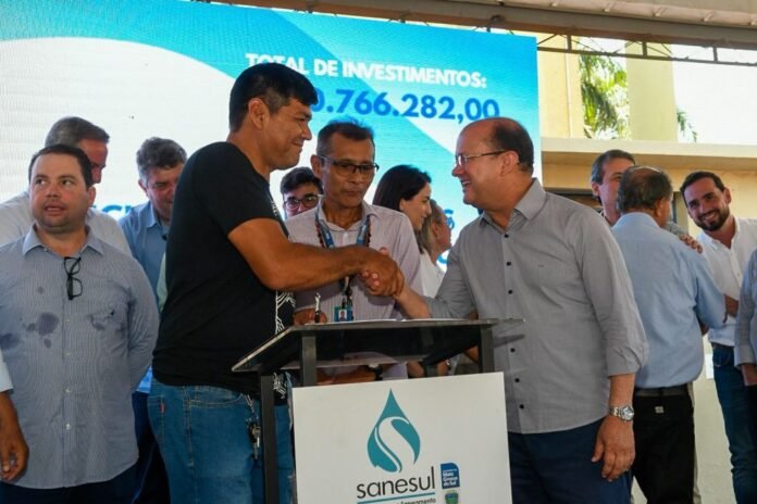 Two men shake hands at a podium during a formal event, with colleagues gathered around and a blue presentation screen in the background showing numbers.
