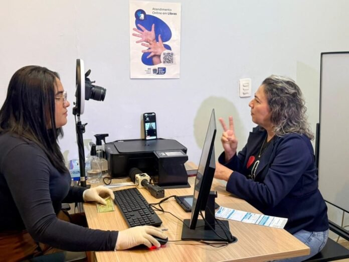 Two women sit across a desk in an office; one works at a computer with a webcam setup, while the other signs with two fingers, with a poster about online Libras service on the wall behind them.