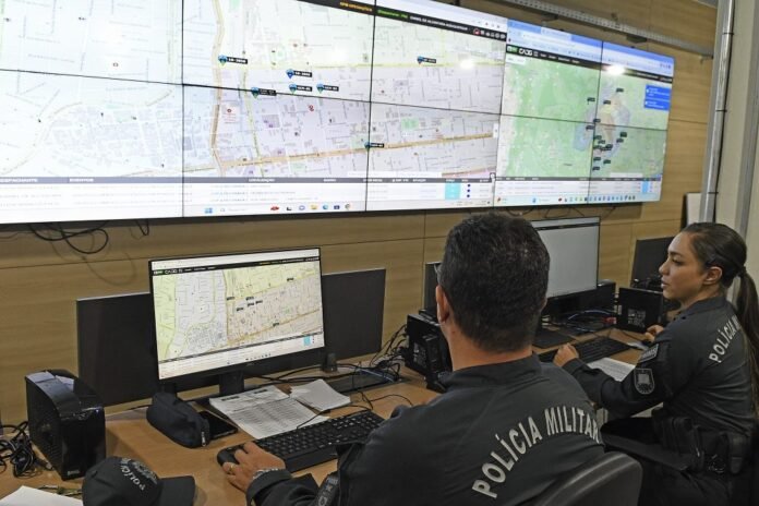 Two Polícia Militar officers monitor a multi-screen map dashboard in a command center.