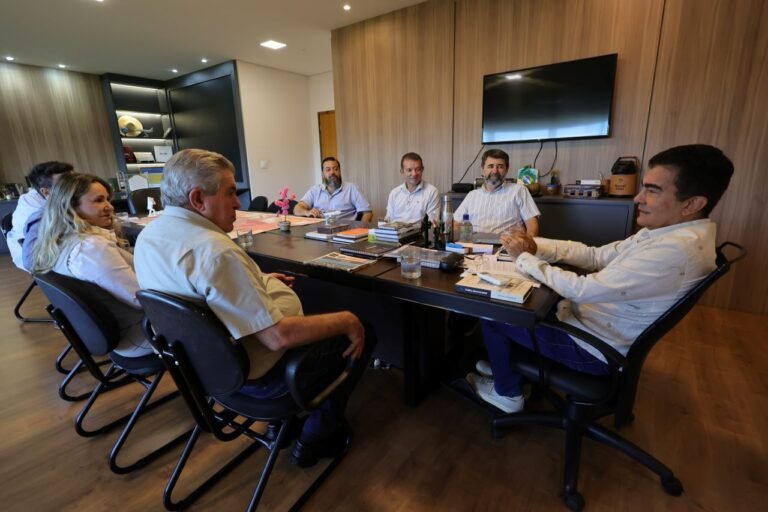 Seven professionals seated around a dark conference table in a modern office, reviewing documents and notebooks during a meeting on a sunny day.