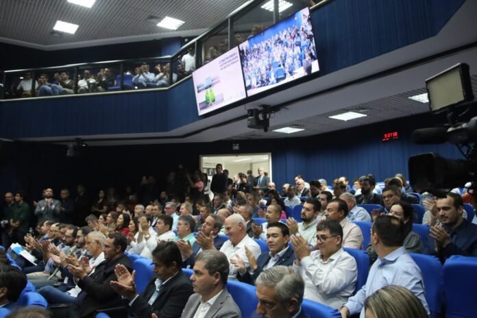 Large conference hall filled with people in blue seats applauding while a camera films the event. Screens above show audience footage.