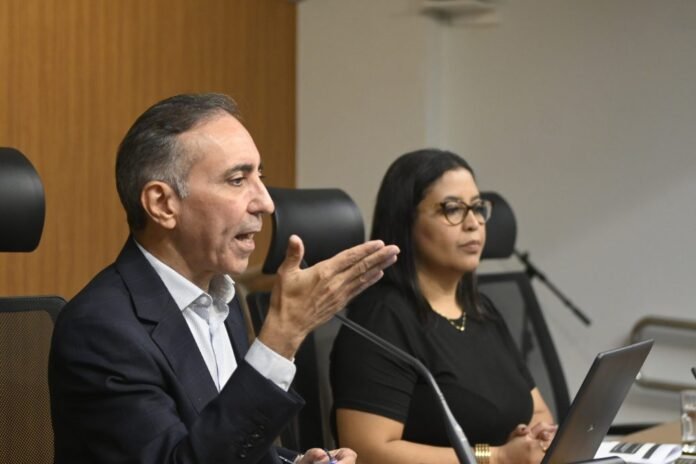 Man in a suit speaks into a microphone at a panel, gesturing with his hand while a woman in black listens beside him.