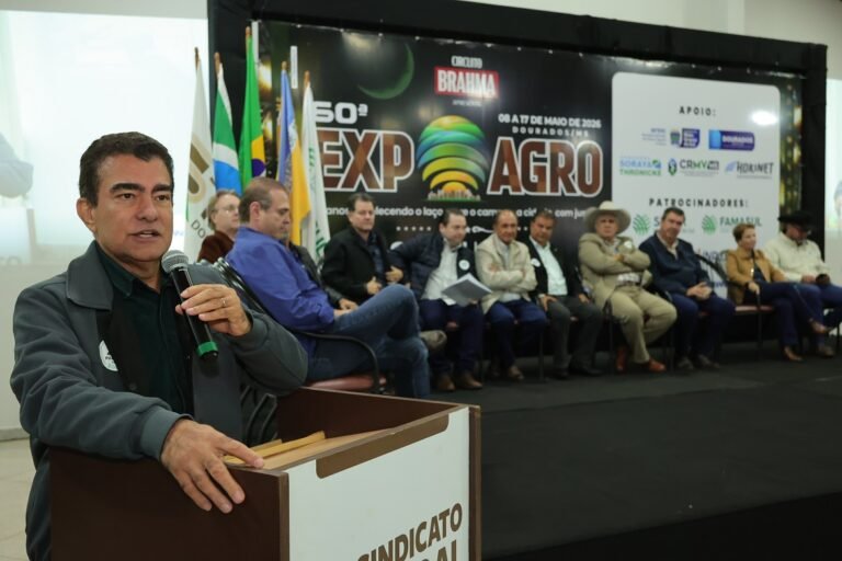 Man at a podium speaks into a microphone while a panel sits on a stage behind him at an Expo Agro event with flags in the background.