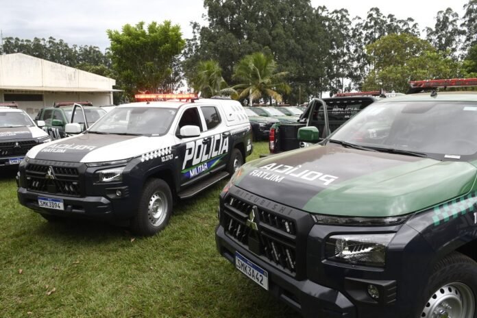 Polícia Militar pickup trucks parked on a grassy field with palm trees in the background and red emergency lights visible.