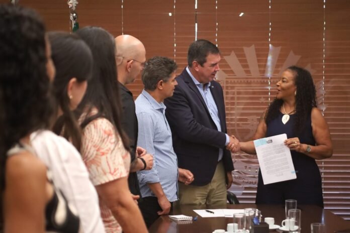 Man in a blazer shakes hands with a woman holding a document at a meeting, while colleagues watch from the side.