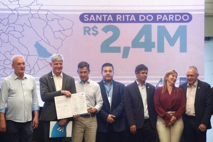 Group of seven people in business attire on a stage; two men in the center hold documents as a large screen reads 'SANTA RITA DO PARDO' and 'R$ 2,4M'.