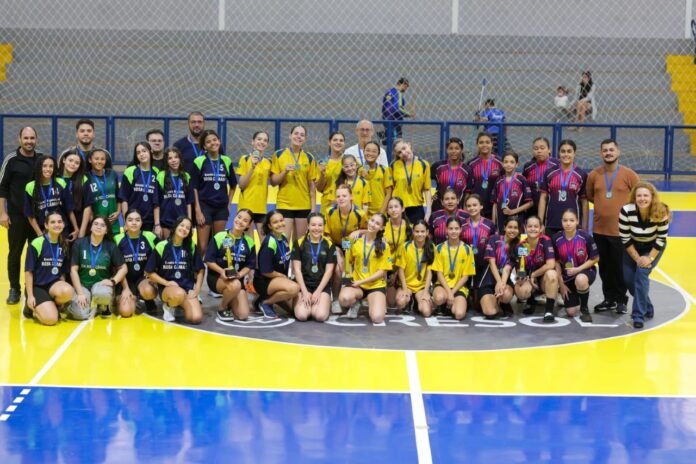 Group photo of youth girls' volleyball teams in colorful uniforms posing with medals on an indoor gym court. Coaches stand with them at the back.