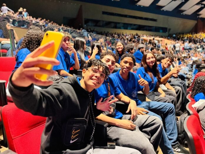 Group of students in blue shirts posing for a selfie in a stadium, a yellow phone held up in the foreground.