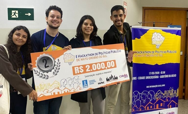 Four young people pose with a large prize check reading R$2.000,00, second place at a Hackathon de Políticas Públicas, backdrop includes event banner.