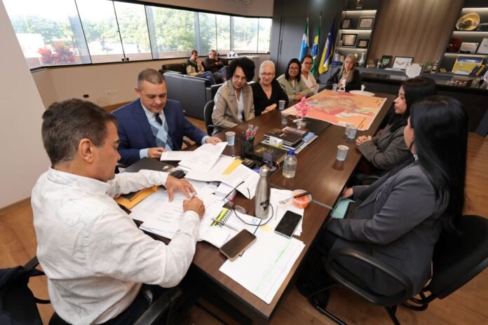 A diverse group of professionals sits around a large conference table in a modern office, reviewing documents; a map, notebooks, and devices are on the table, with flags and windows in the background.