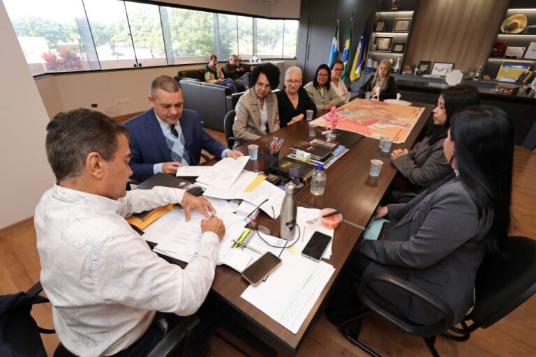 A diverse group of professionals sits around a large conference table in a modern office, reviewing documents; a map, notebooks, and devices are on the table, with flags and windows in the background.