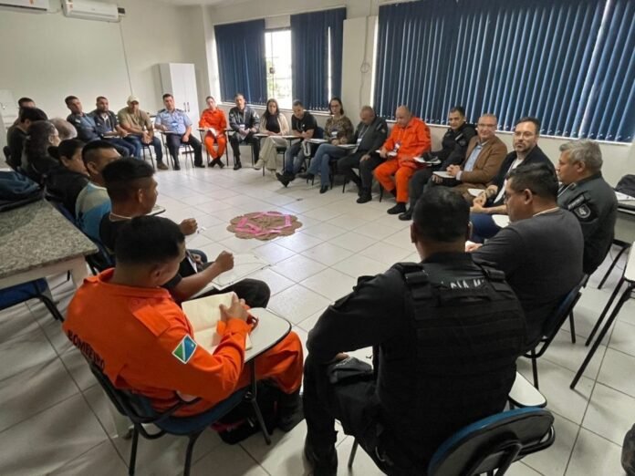 Group of about 20 people seated in a circle in a meeting room; several in orange jumpsuits and others in uniforms, taking notes under bright windows.