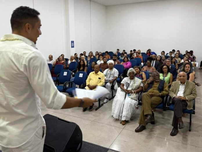 A man in a white shirt speaking to a diverse audience seated in blue chairs in a classroom or lecture hall.