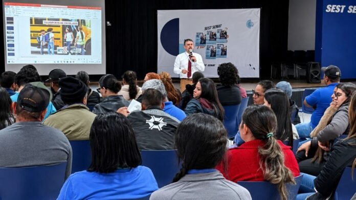 Presenter in a white shirt and red tie speaks into a microphone on stage while a PowerPoint slide about school transport is projected; audience seated in blue chairs.