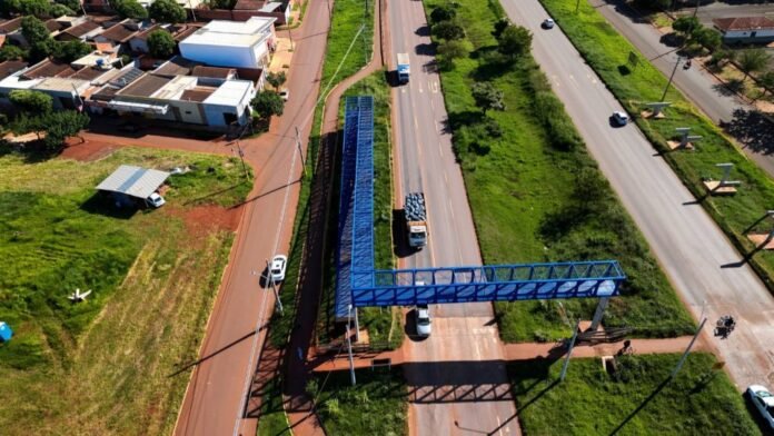 Aerial view of a blue road sign gantry spanning a multi-lane highway with cars and trucks, flanked by a green median and buildings nearby.