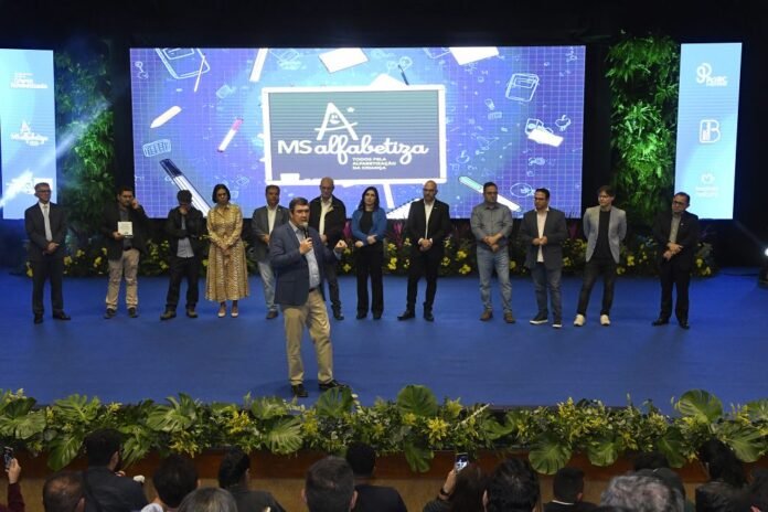 Speaker in a blue blazer addresses the audience on a blue stage, with a line of people and a large screen reading 'MS alfabetiza' behind them.
