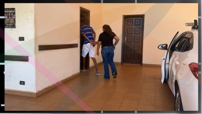 Two people walk toward an open doorway in a beige tiled hallway, with a white car parked to the right.