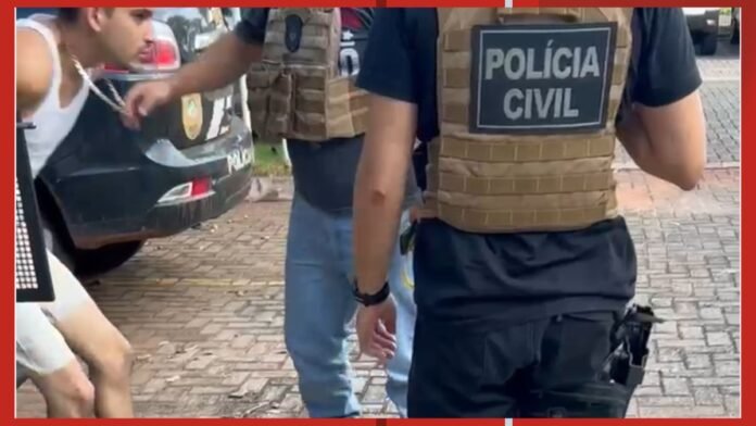 Police officer in tactical vest labeled POLÍCIA CIVIL stands beside a car while a man in a white sleeveless shirt is restrained nearby on a cobblestone street.
