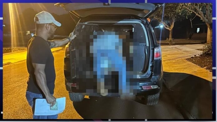 Man in a cap and black shirt stands by the open trunk of a dark SUV at night, a pixelated figure is inside the cargo area.
