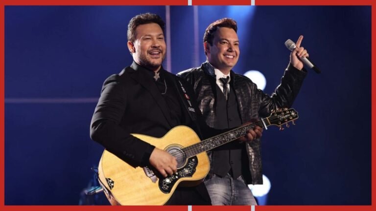 Two male musicians on stage: one plays an acoustic guitar while the other holds a microphone and smiles under blue lighting.