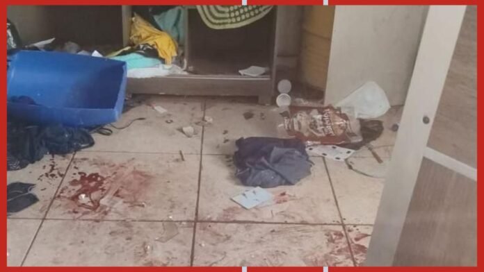 Messy tiled floor with red blood stains, scattered papers and clothing, and a tipped blue plastic bin in a cluttered room.